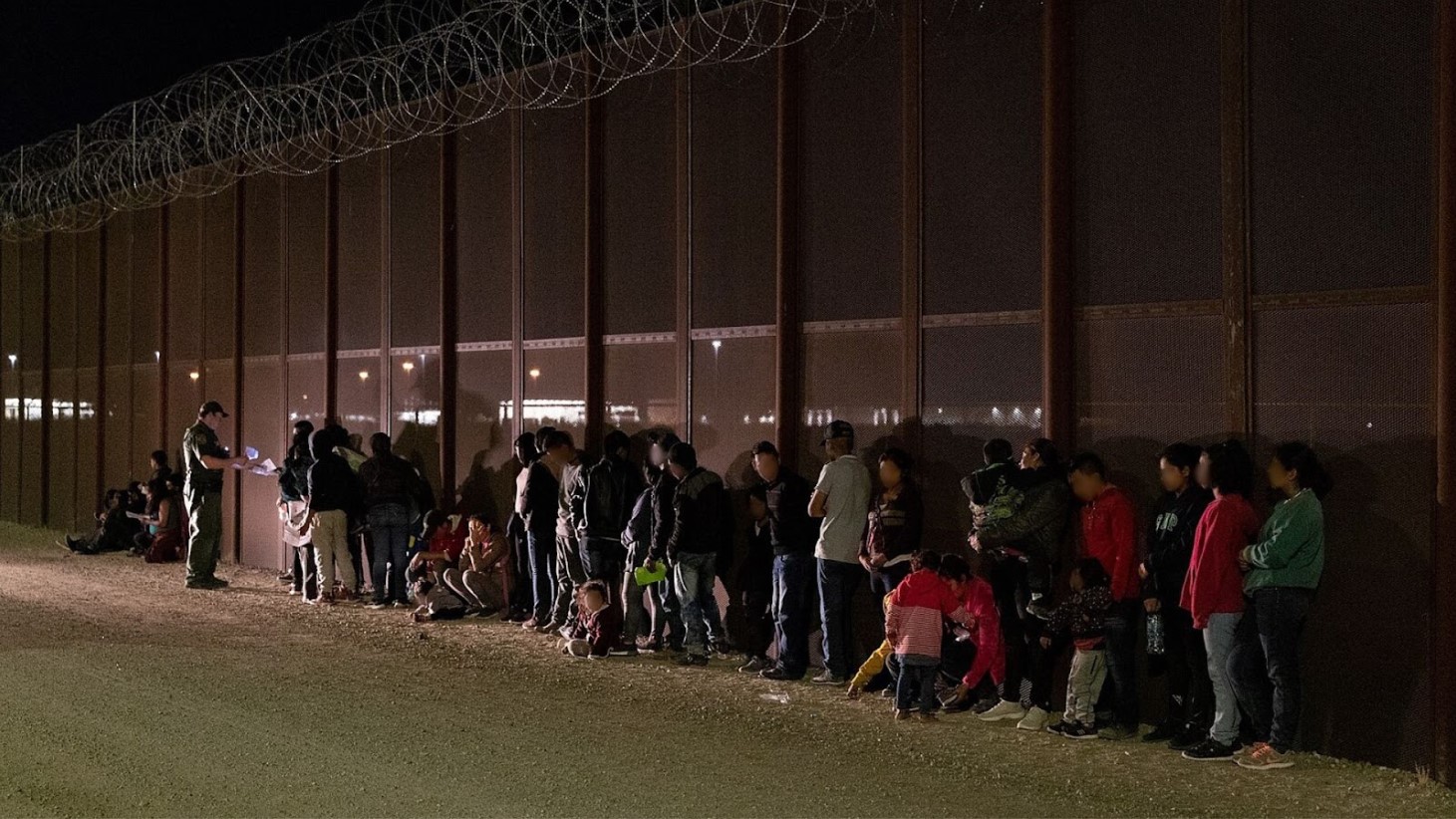 A group of people lining up next to a border fence at nighttime