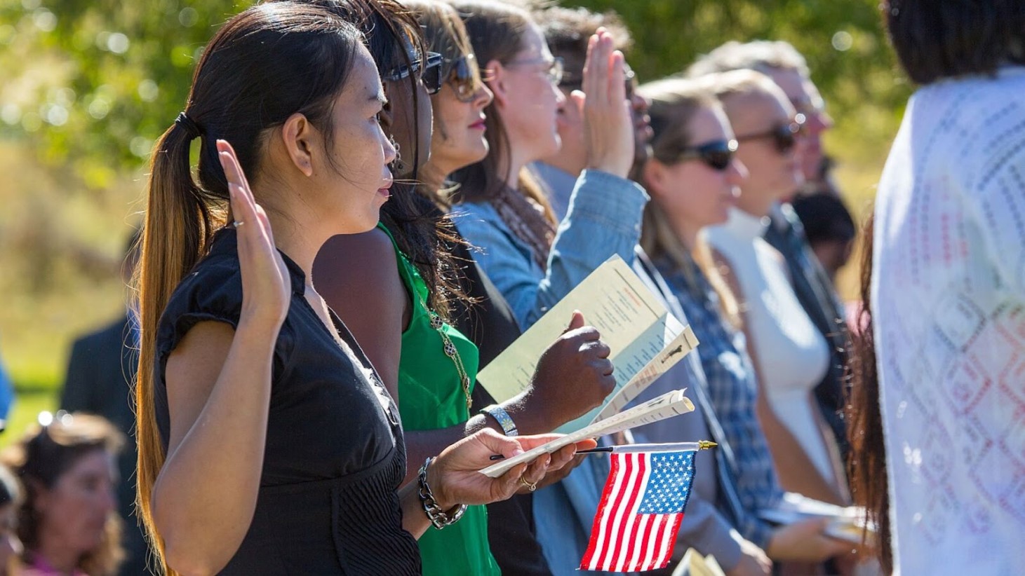 A group of individuals standing outdoors, taking the oath of allegiance in a naturalization ceremony, with each person raising their right hand and some holding small American flags and certificates