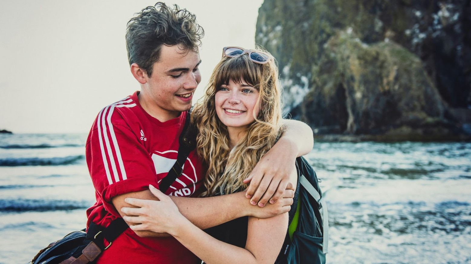 A young couple embraces on a beach. The boy is wearing a red soccer jersey and the girl has long blond hair and sunglasses on her head