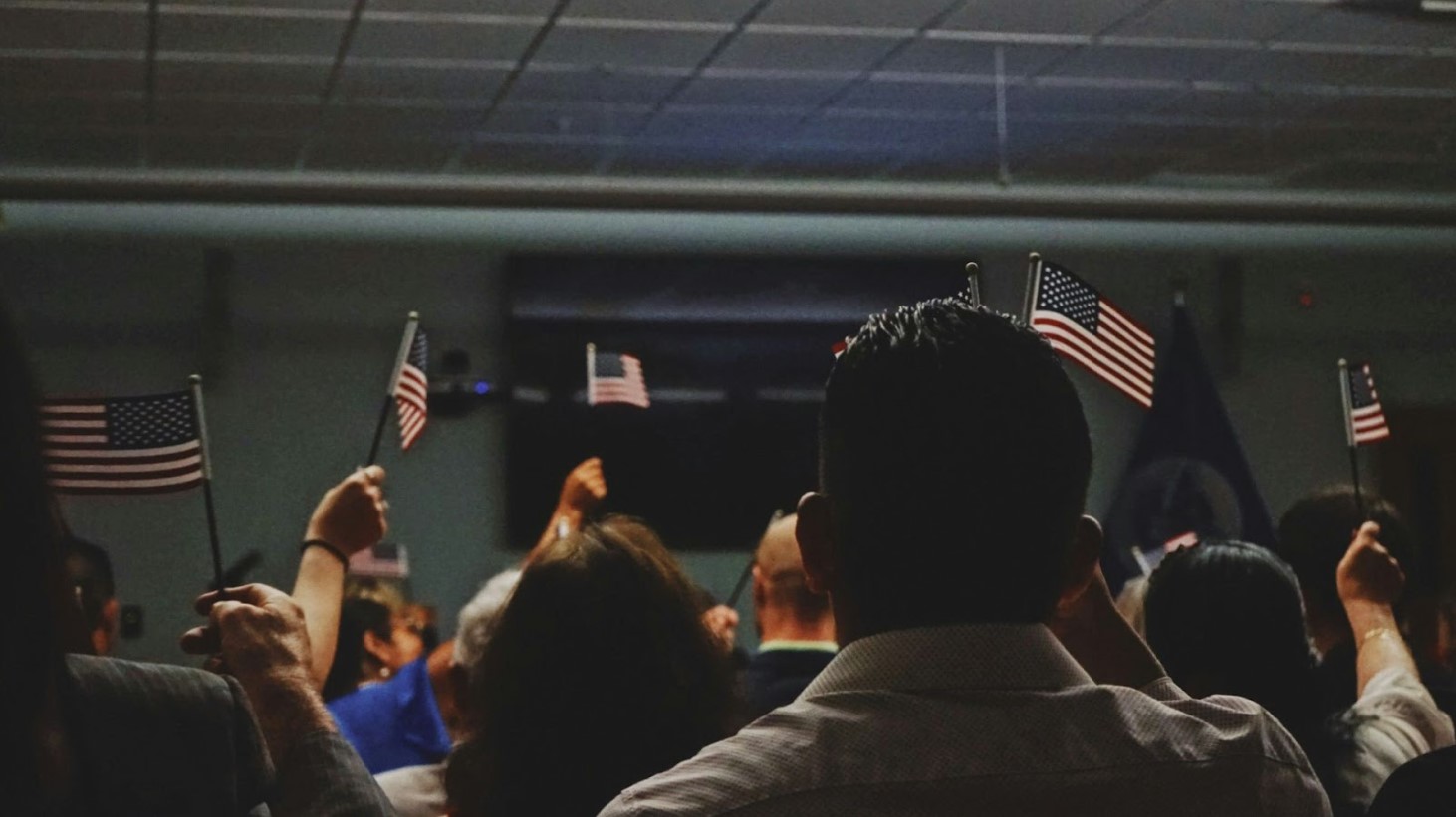 A group of people seen from behind, raising their right hands and holding small American flags during a citizenship oath ceremony