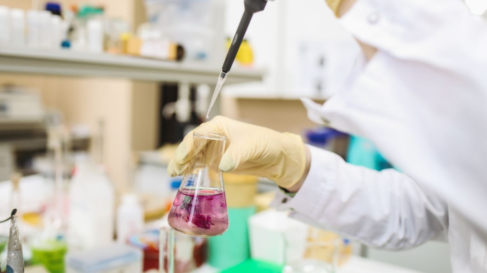 A scientist holds a beaker with purple liquid and a pipet while wearing yellow gloves and a white coat