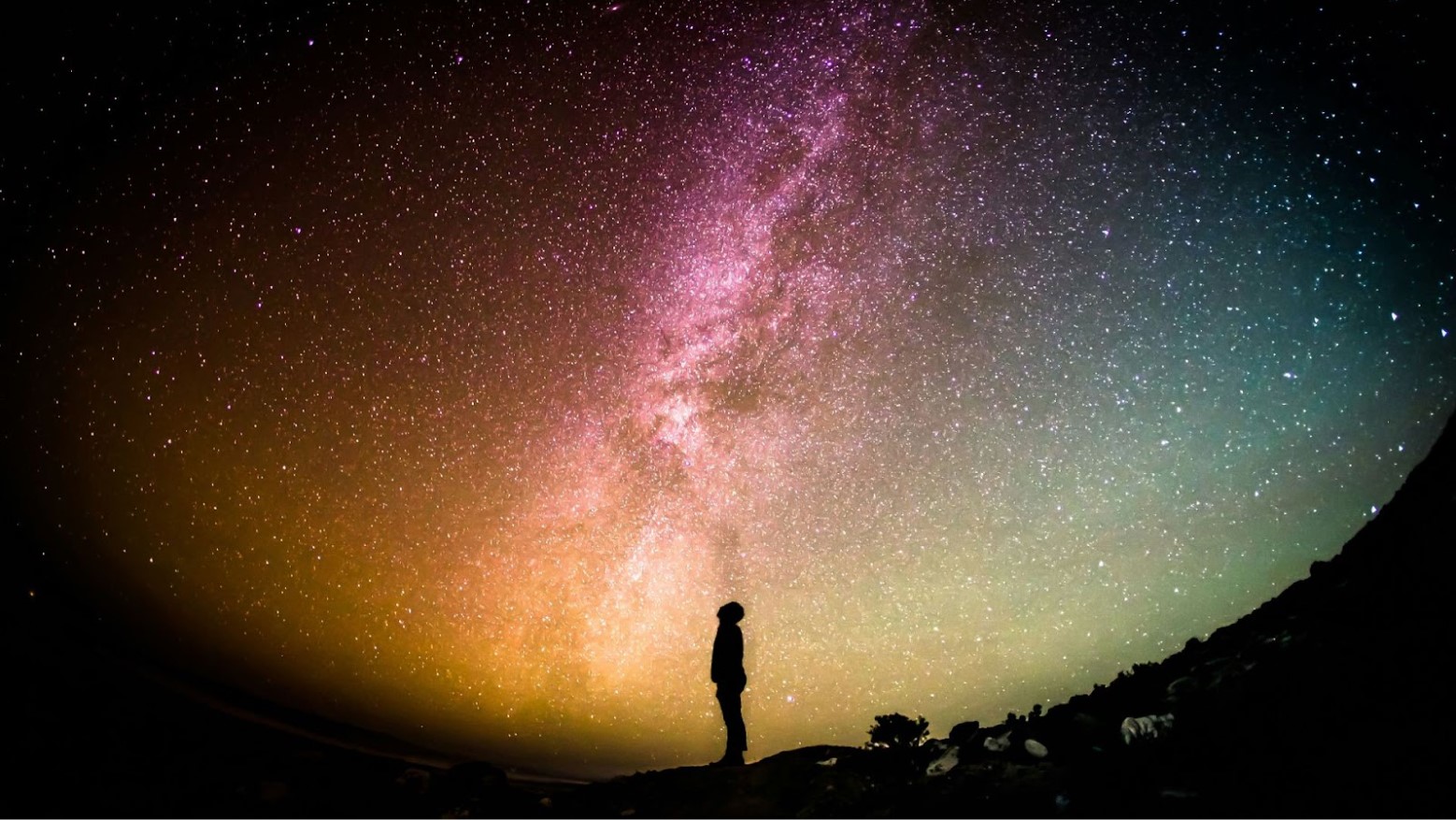 A silhouette of a person standing underneath a colorful starry night sky.