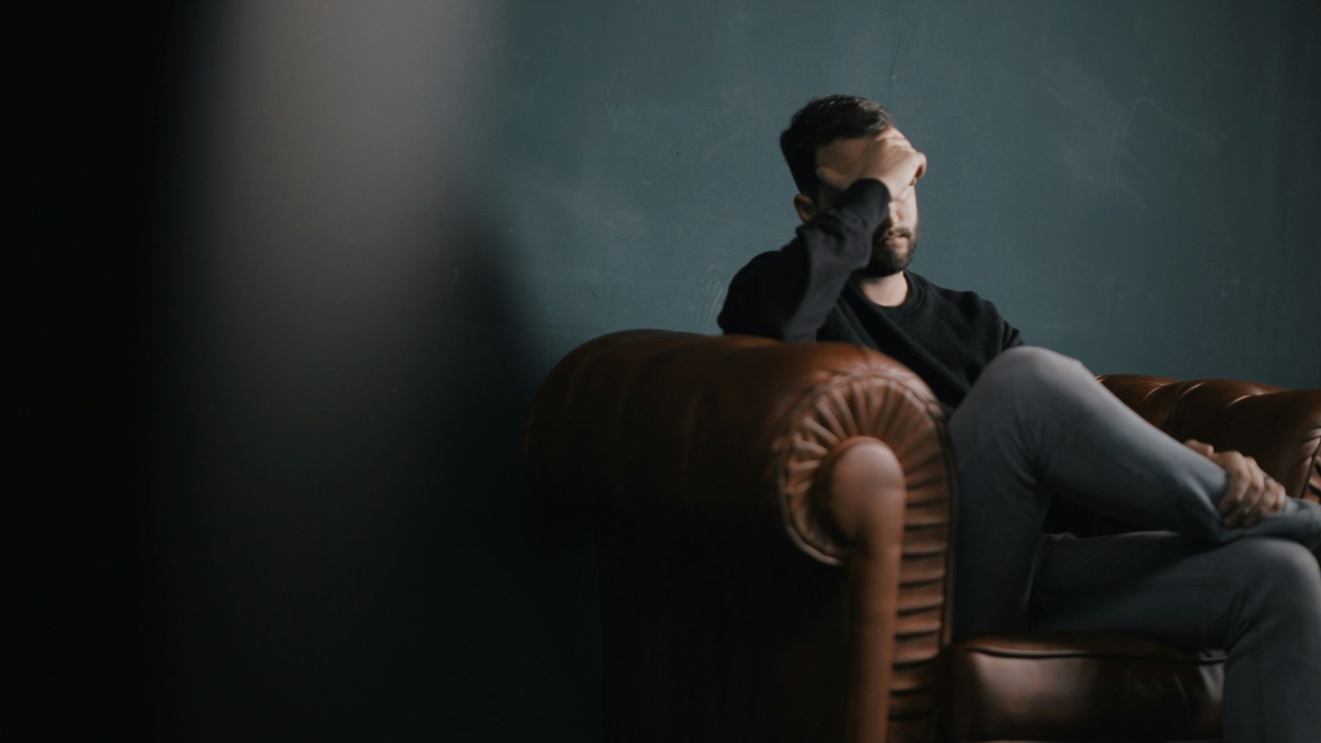 A worried man sitting on a leather couch with his hand on his head.
