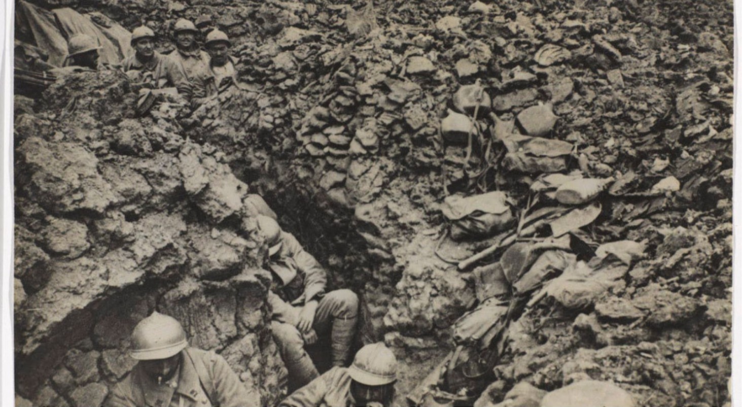 Black and white photo of World War I soldiers huddled in a deep trench. They wear helmets and look upward