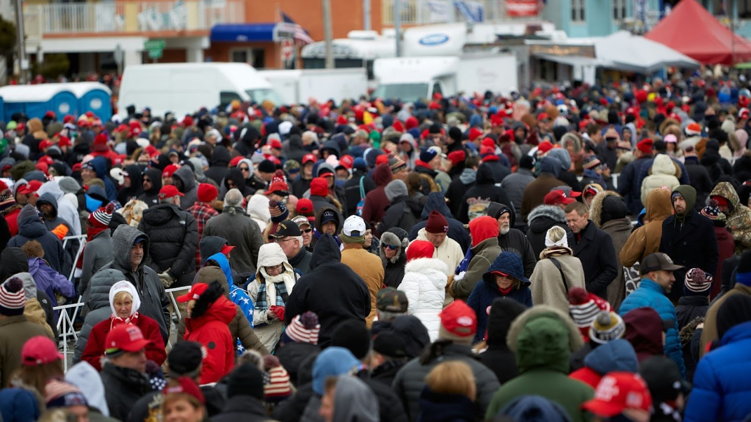 A large crowd of individuals bundled up in cold-weather clothing, many wearing hats in various colors, gathering closely in an outdoor setting