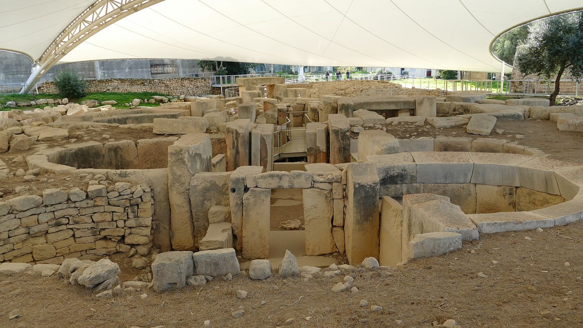 A photo of the Tarxien Temples archaeological site in Malta. Visible are the exposed foundational remains and partial reconstructions of ancient structures. The site is a complex of stone walls, steps, and chambers made from large limestone blocks. A modern canopy stretches overhead to protect the ruins