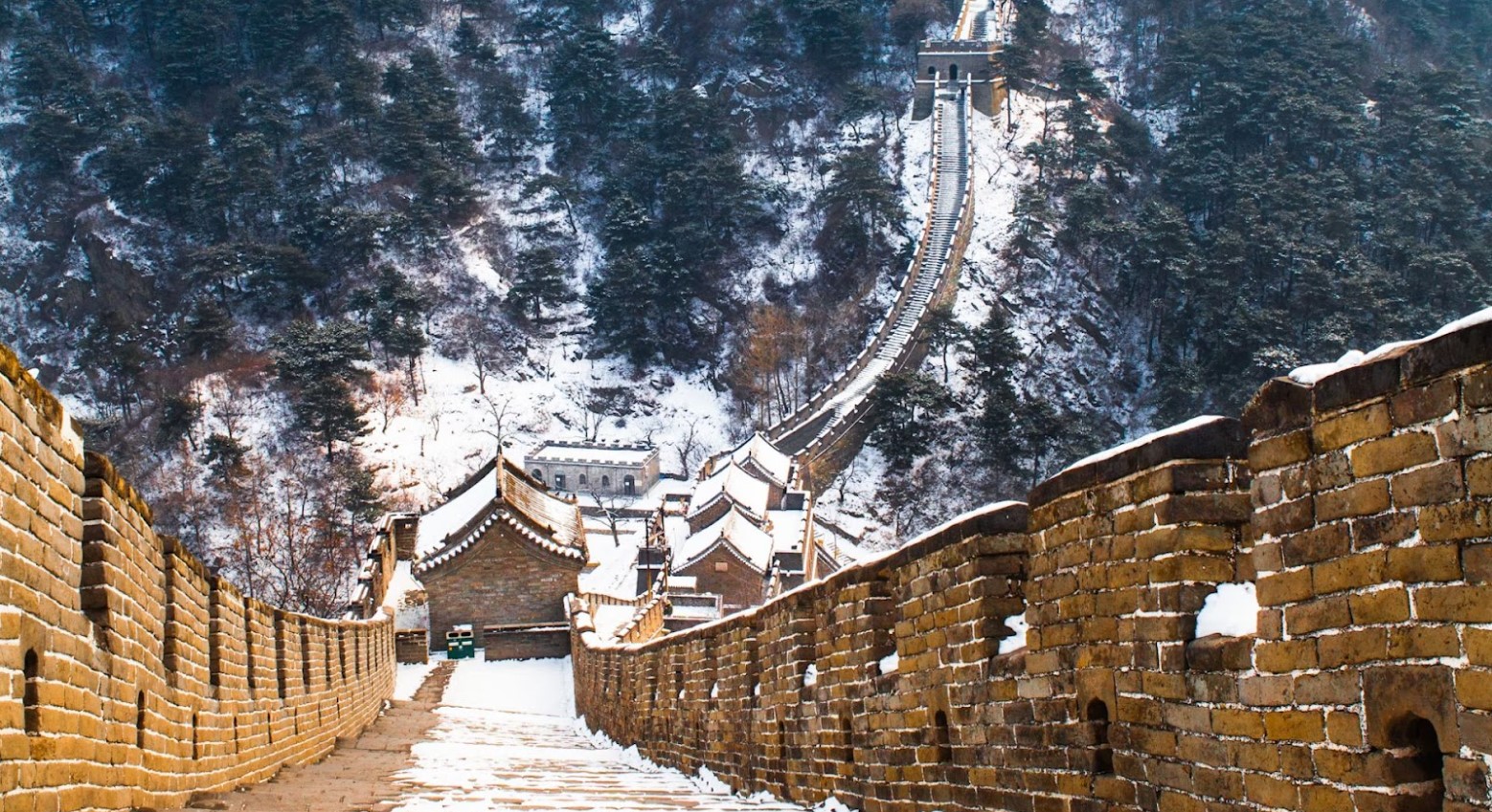The Great Wall of China during winter. There is snow on the steps of the wall. The wall goes down and then back up the hill.