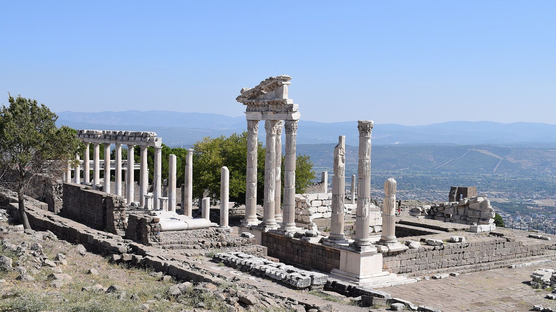 Photograph of the ruins of an ancient classical temple situated on a hilltop, with a clear blue sky above and a panoramic view of the valley below. The temple consists of standing and fallen columns, some with Corinthian capitals