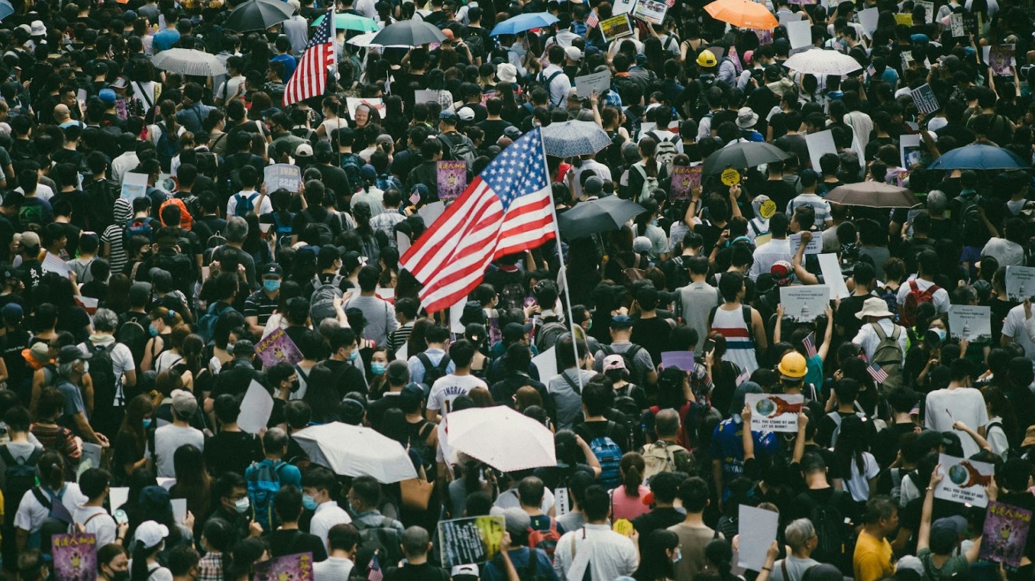 A dense crowd of protesters, many holding umbrellas and signs, with an American flag being held high above the sea of people