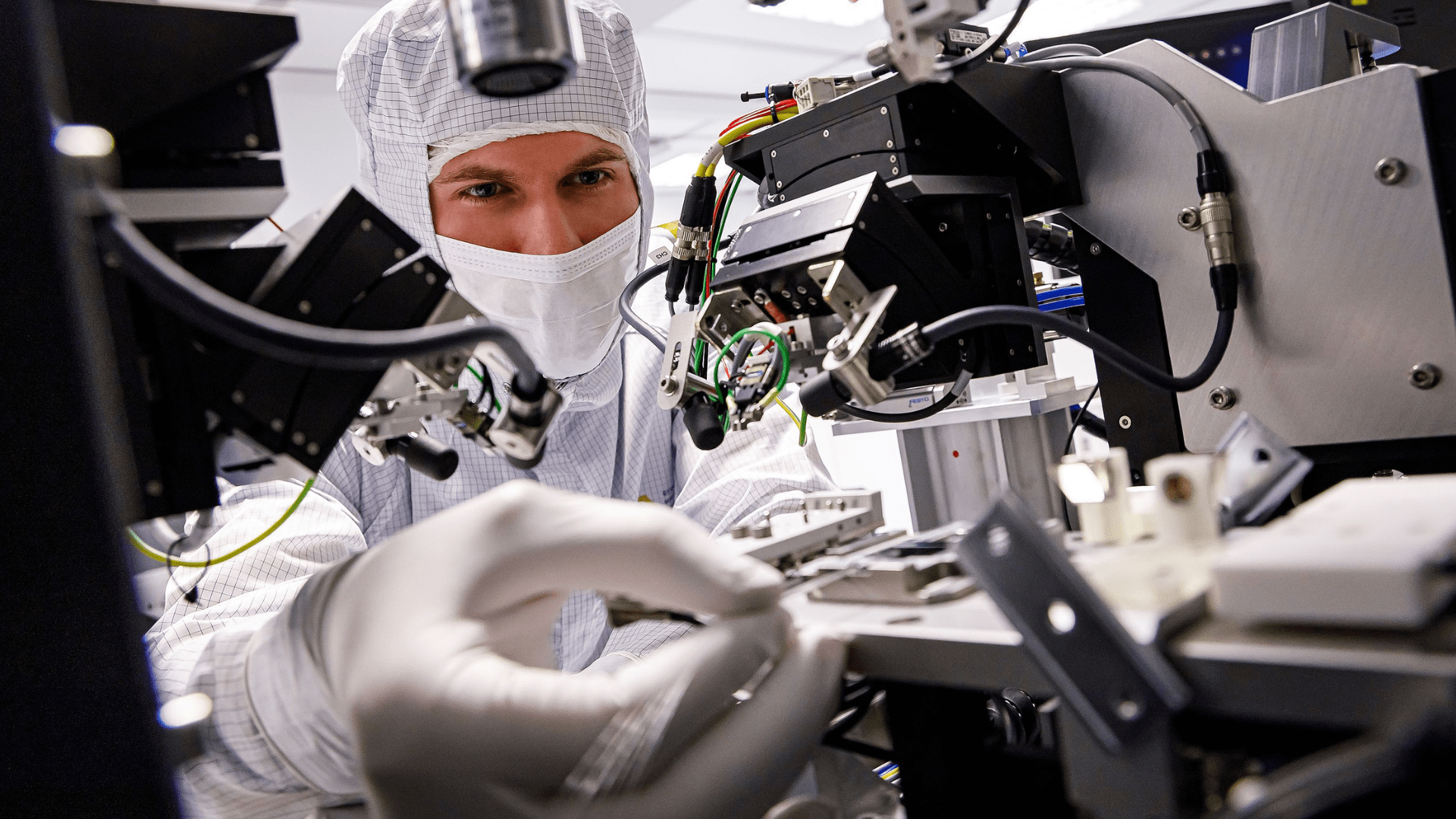 A scientist wearing scrubs, a mask, and gloves handles laboratory equipment.