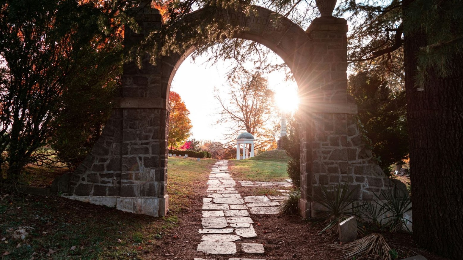 A cemetery seen through a brick archway, with grass, trees, and sunlight seen through the arches.