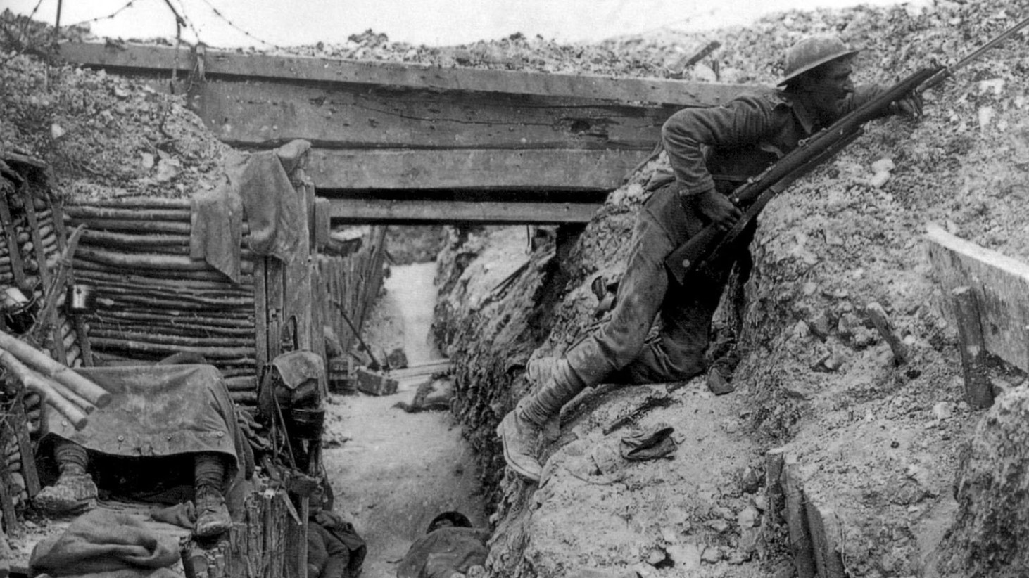 Black and white photograph of a World War I soldier peering over the edge of a trench with his rifle at the ready. Another soldier lies in the foreground, seemingly resting or wounded