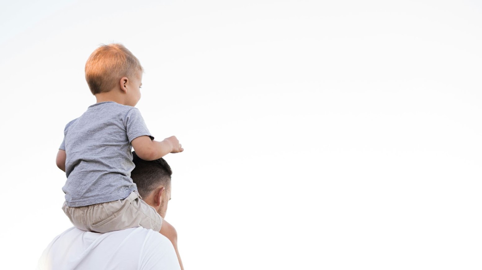 A father carrying his child on his shoulders in the daytime against a clear white sky and background.