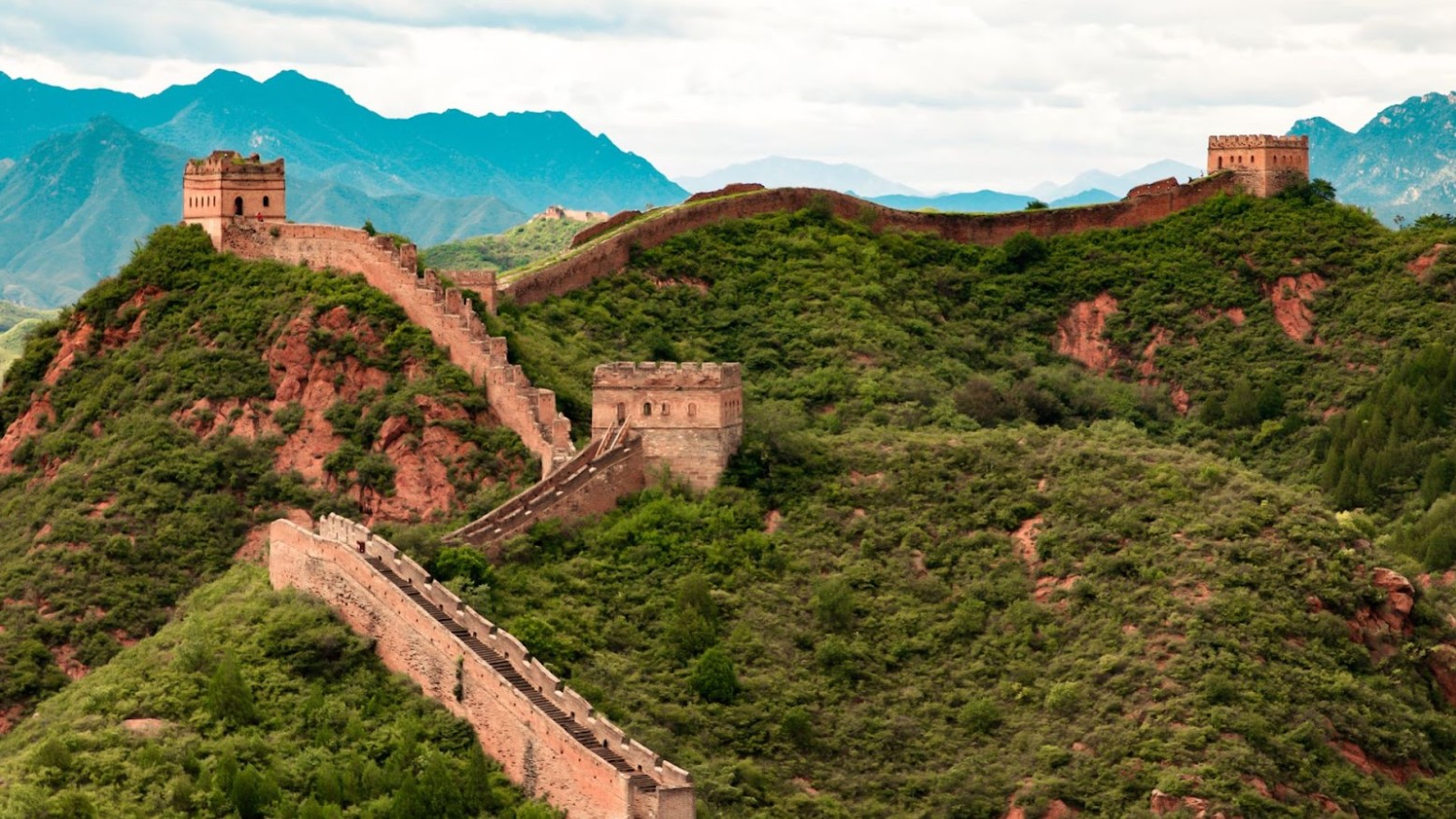An image of the Great Wall of China. As the wall is going upwards it curves round to the right. The wall is on a hill and there are hills behind it.