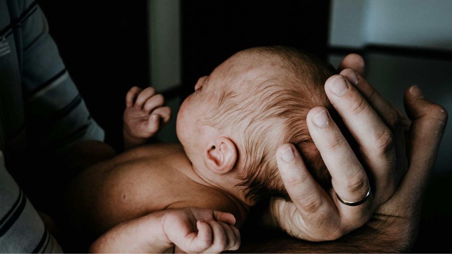 A newborn baby cradled gently in a father's hands, which are securely supporting the baby's head and body