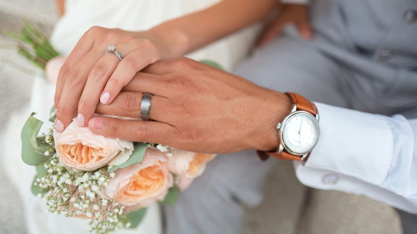 A close-up of a bride and groom's hands together, with the bride's engagement ring and the groom's wedding band visible