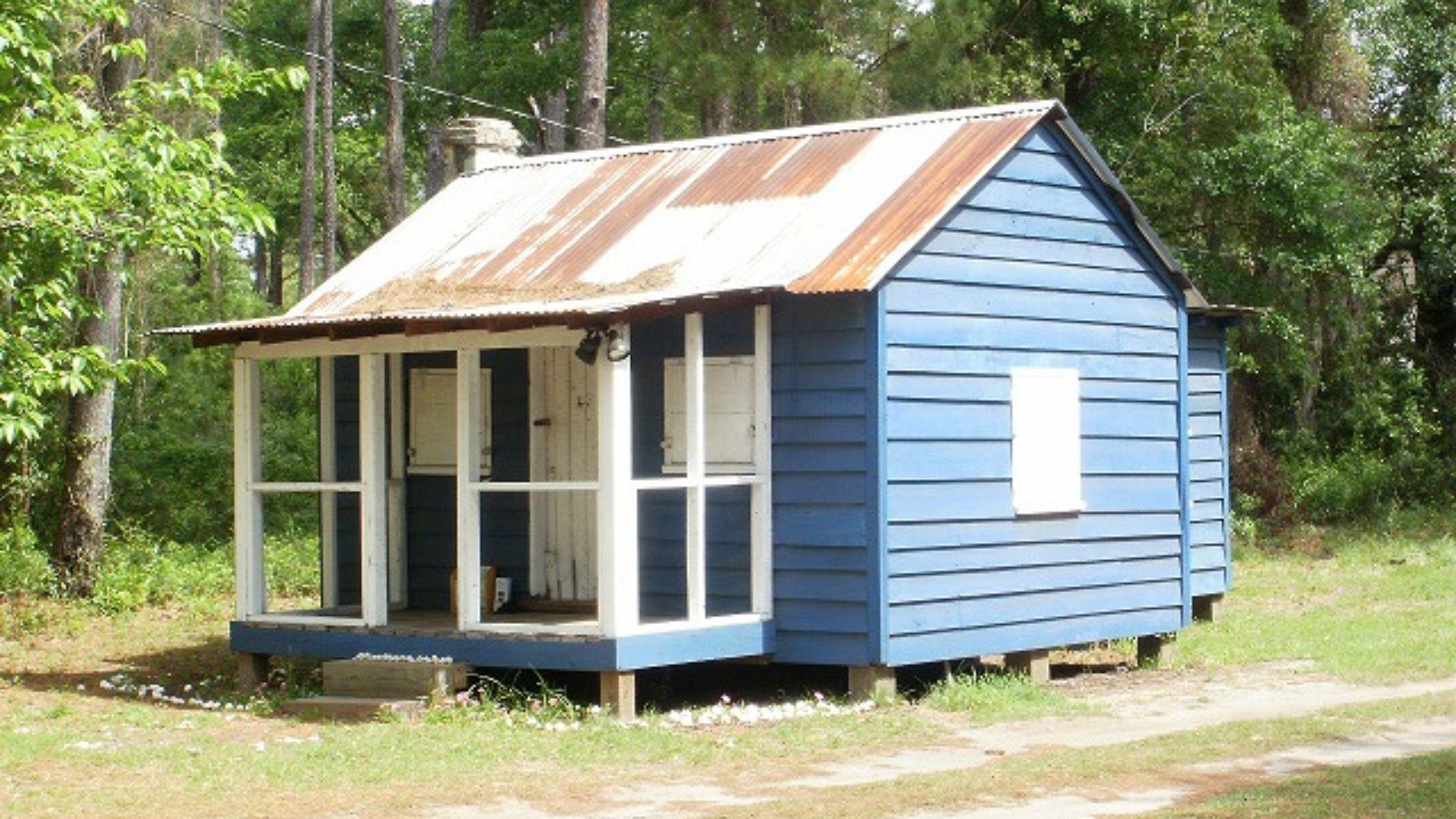 A small, single-story wooden house painted in blue with white trim, featuring a corrugated metal roof, a front porch with steps, and a single white-shuttered window