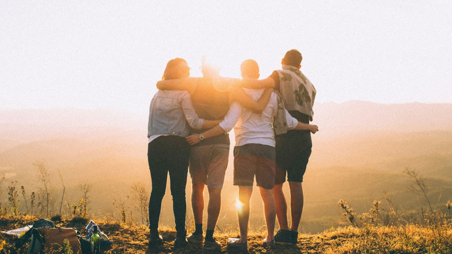 Four friends stand closely together, embracing shoulder to shoulder on a hilltop, backlit by the glowing sun setting on the horizon