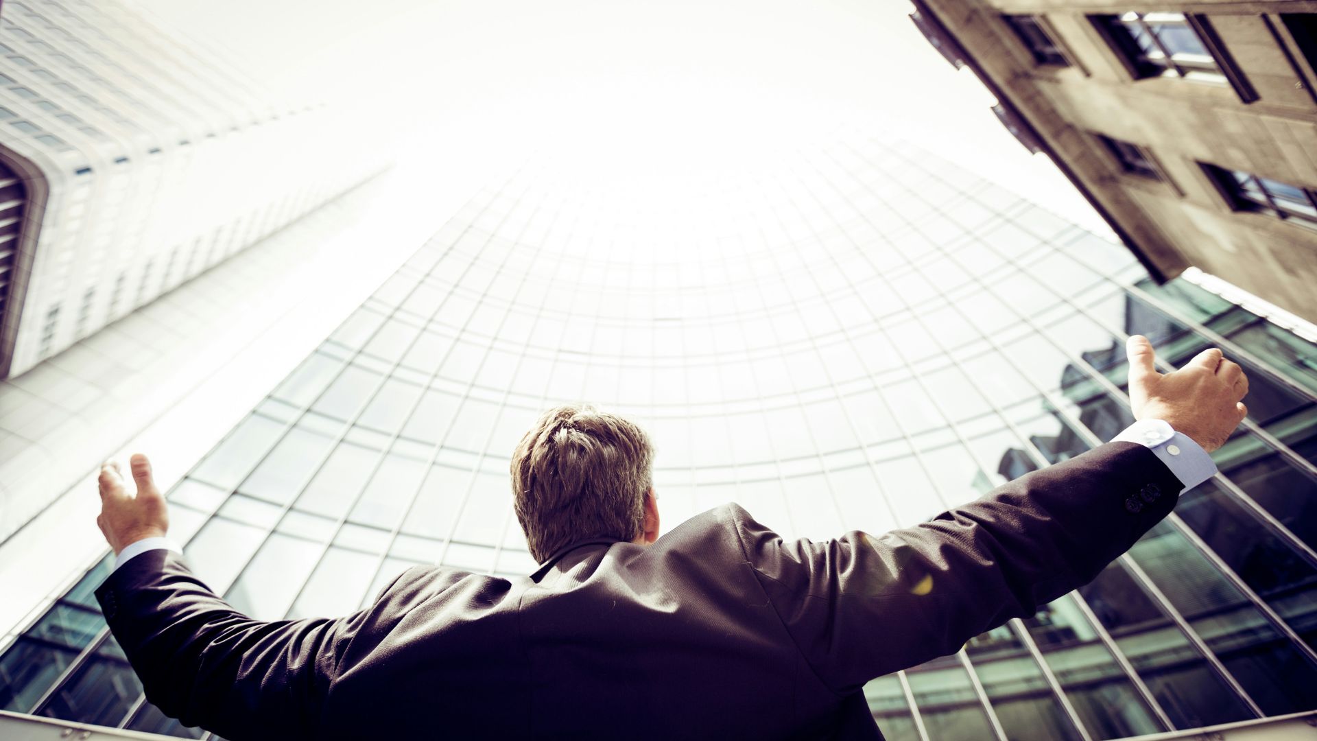A man in a suit raising his arms to the sky in front of tall buildings.
