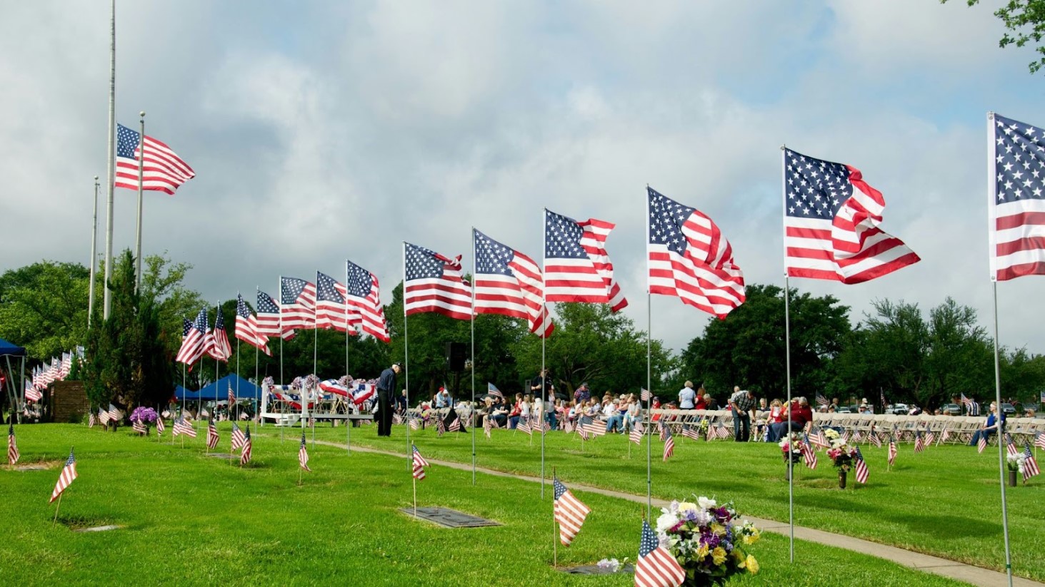 American flags fluttering in the wind at a Memorial Day event, with people seated in chairs