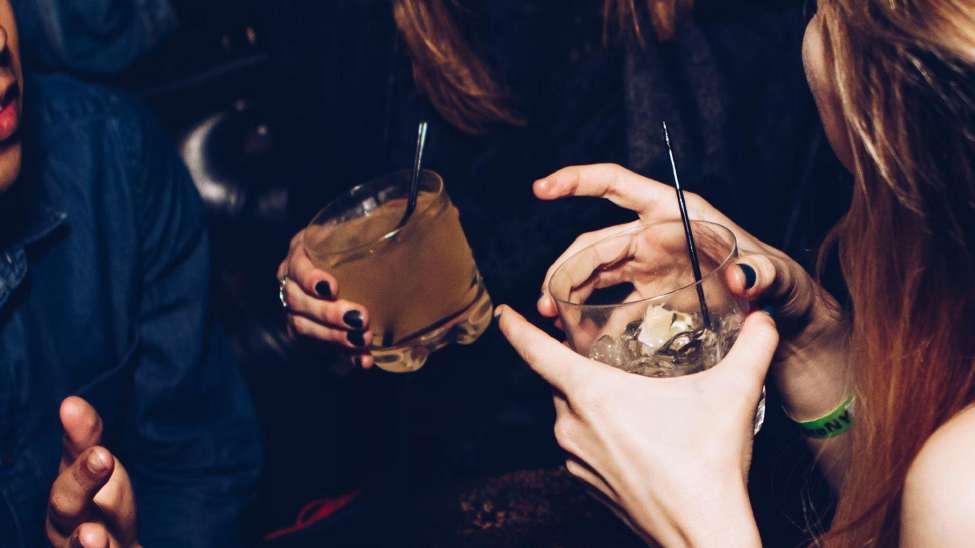 A group of people sat down to drink alcohol. The two girls in the picture have a drink in a tumbler glass with a black straw in both glasses.