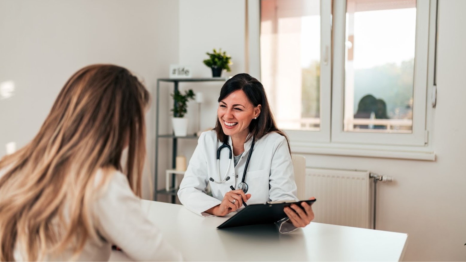 A young woman sits with her back to the camera while a doctor smiles and makes notes on her chart