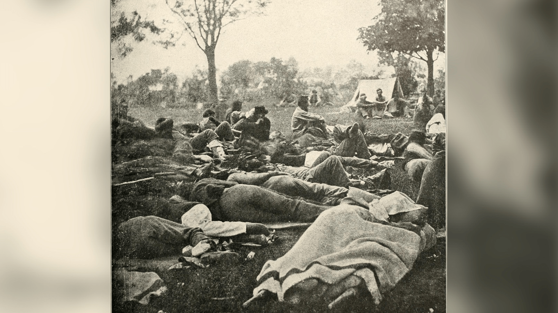 Black-and-white photo of injured people lying under blankets in a Civil War battlefield campground.