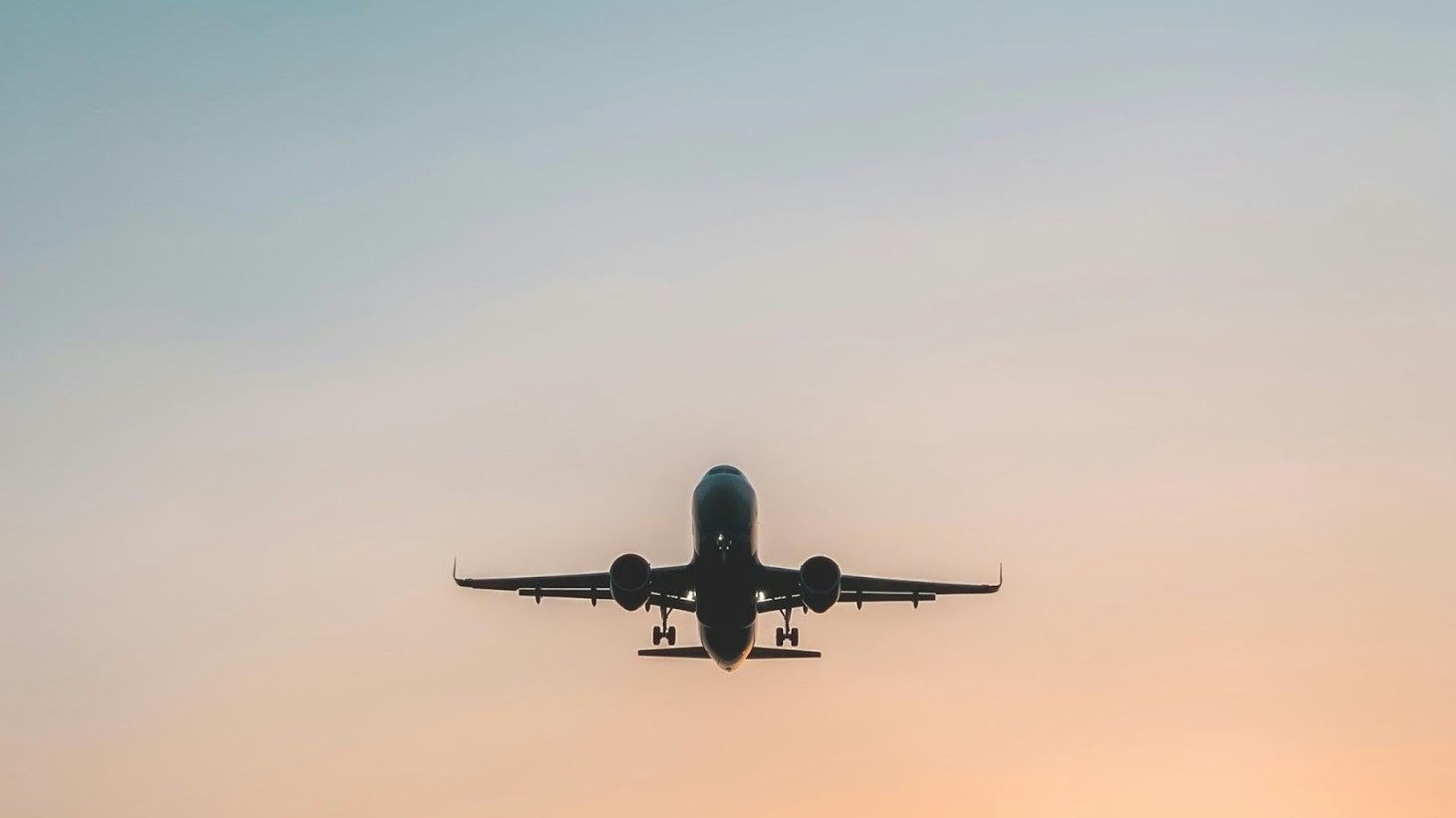 An airplane captured in the center of the frame against a soft sunset sky, with the aircraft's landing gear down as it prepares for landing