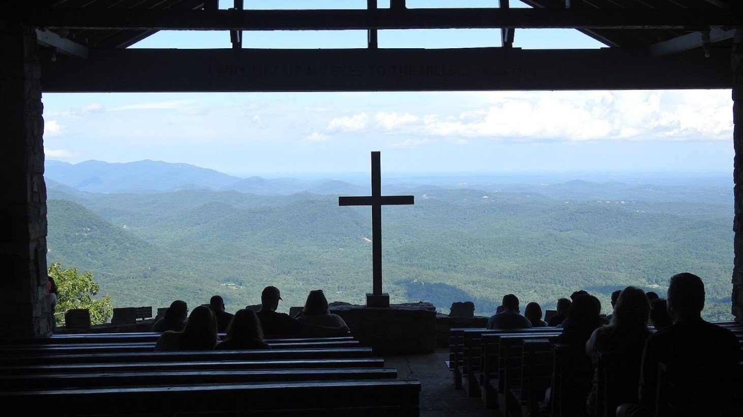 Silhouettes of people sitting on wooden benches in a chapel with open walls, facing a large cross in the foreground that overlooks a panoramic view of mountains