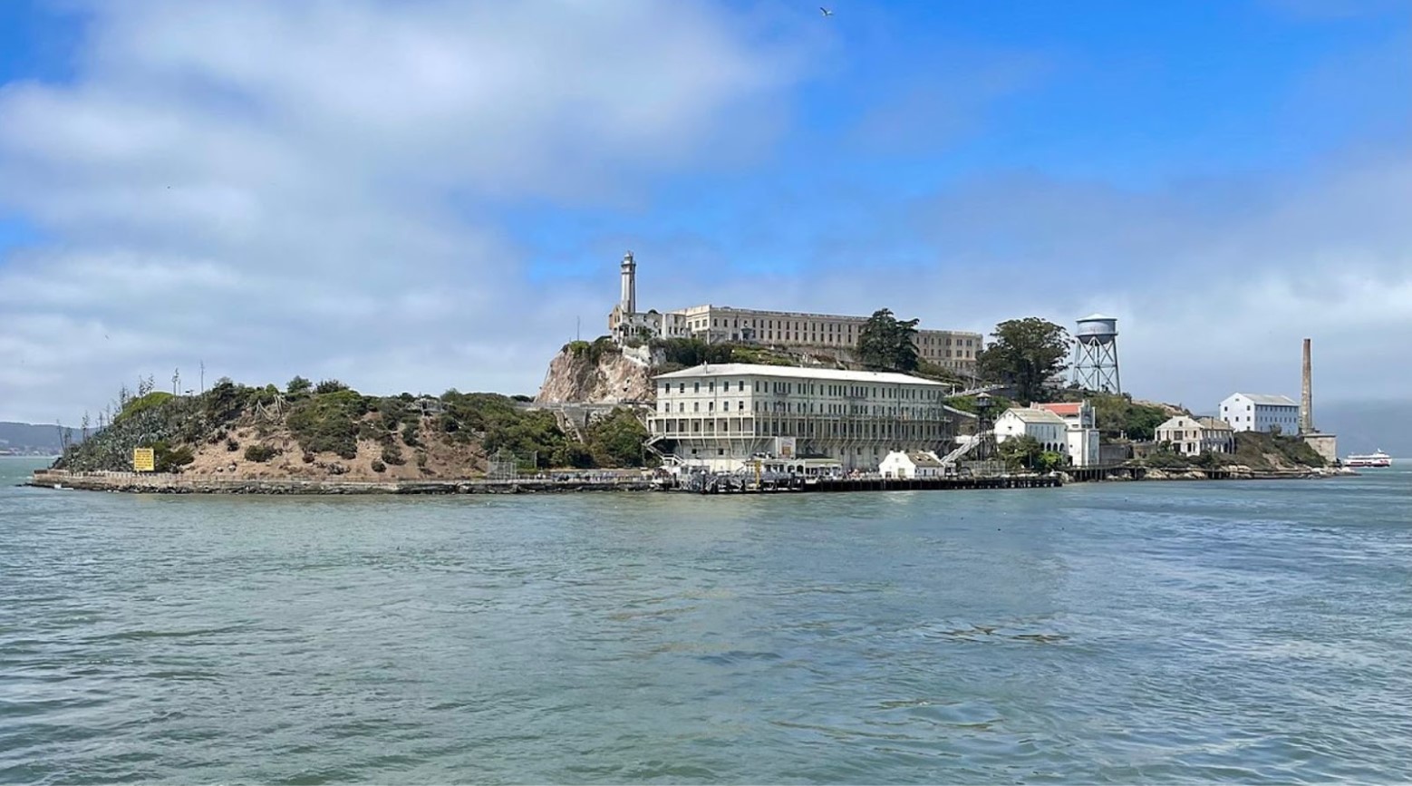 An image of Alcatraz Island. There are various buildings on the island, including the prison, and it is surrounded by the sea.
