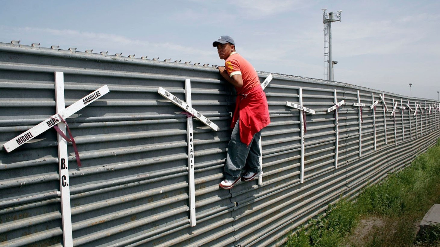 A person stands next to a border fence adorned with crosses bearing names, each cross commemorating individuals who have died at the border