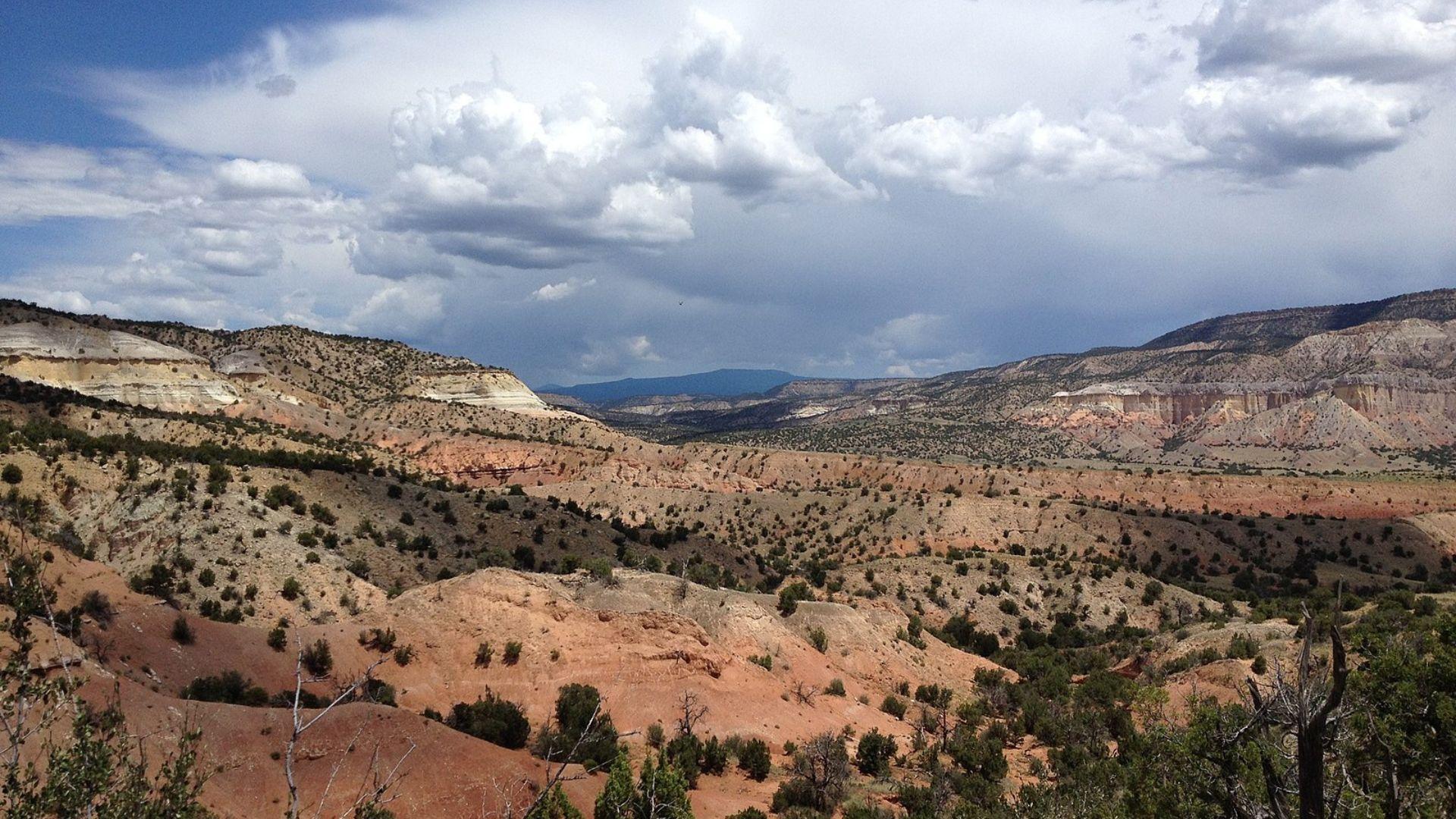 A panoramic view of a desert landscape featuring layered plateaus with varying shades of red, white, and brown under a partly cloudy sky