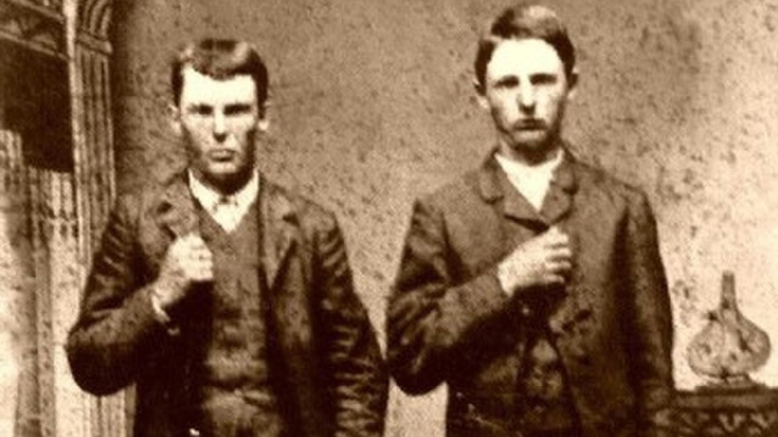 A black and white photo of Frank and Jesse James wearing suits and holding the jacket with their hand.