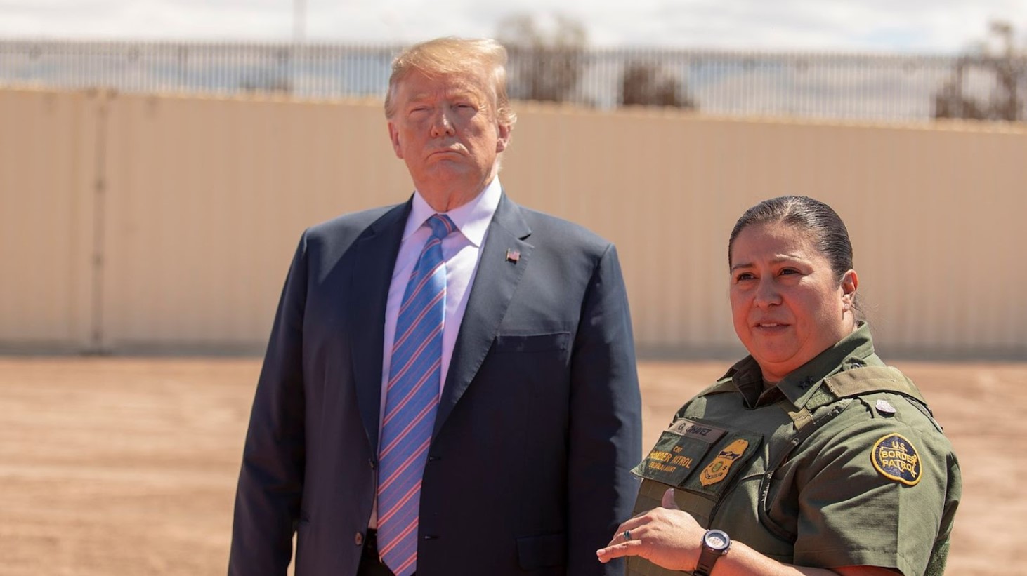 Donald Trump standing next to a border patrol officer in uniform, in front of a tall metal fence under a clear sky