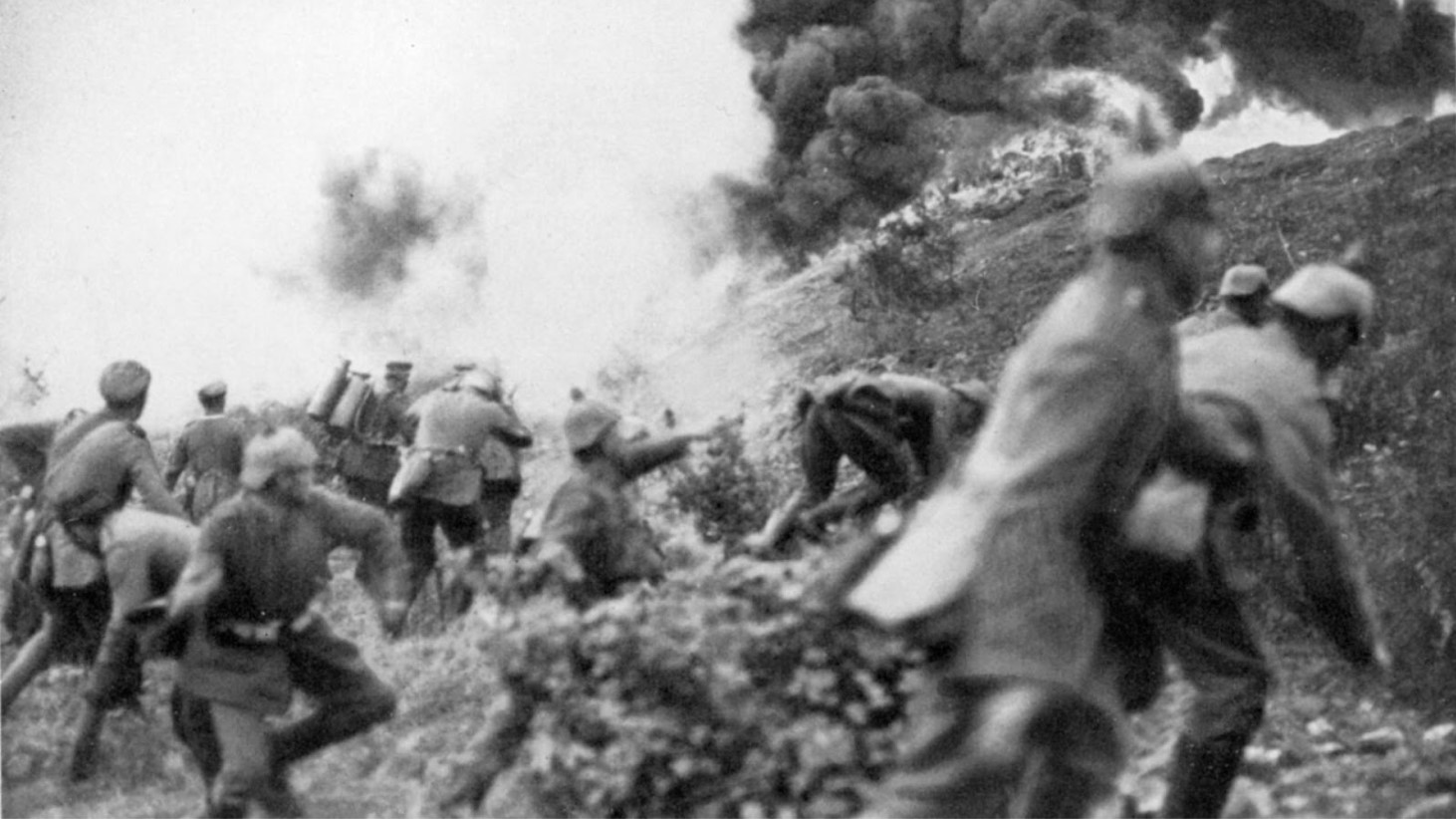 A grainy black and white photograph captures a group of World War I soldiers advancing uphill under fire. An explosion is seen in the background