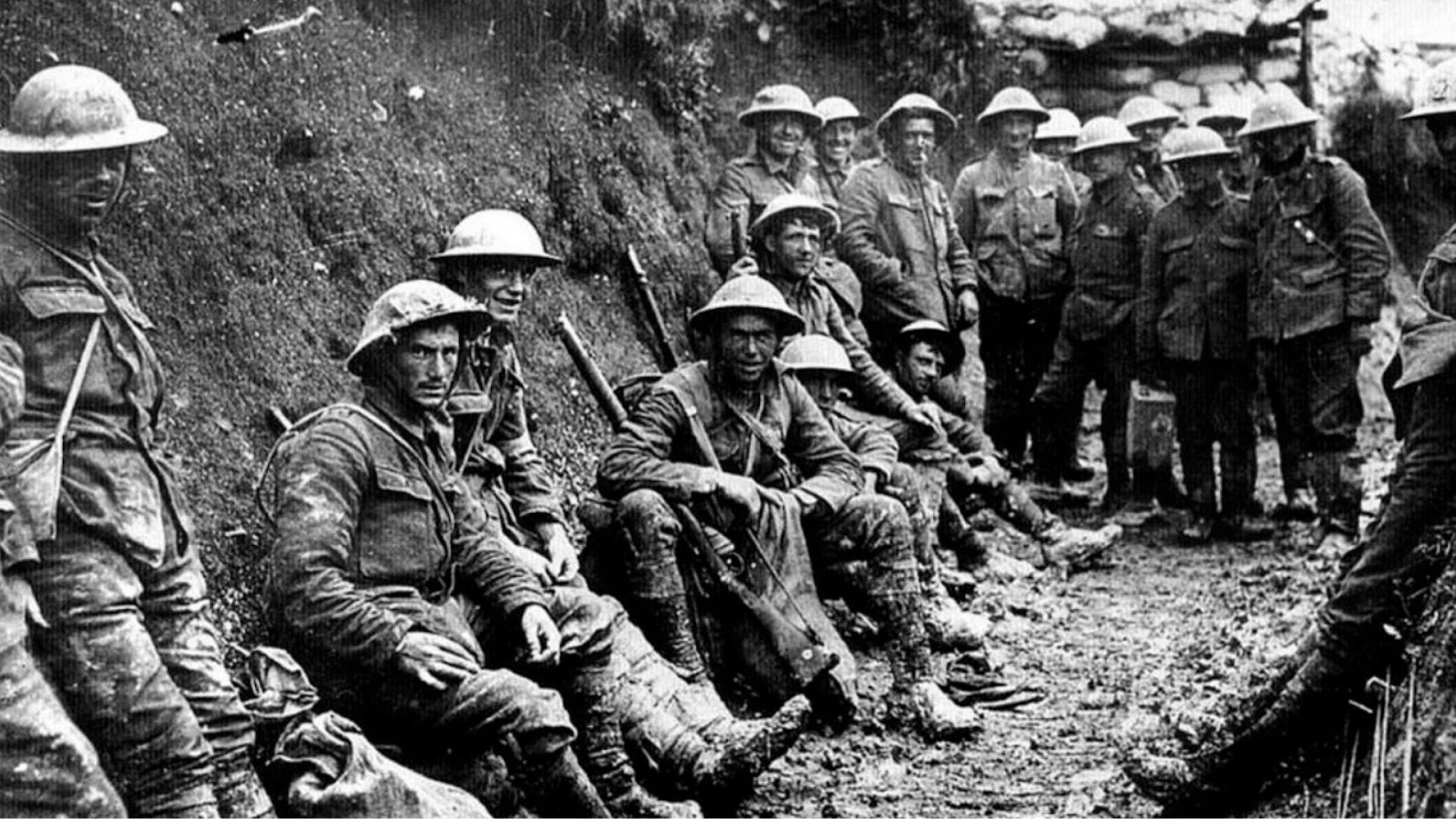 A black and white historical photograph showing a group of World War I soldiers inside a trench. They are wearing helmets and mud-covered uniforms, with rifles in hand or resting beside them