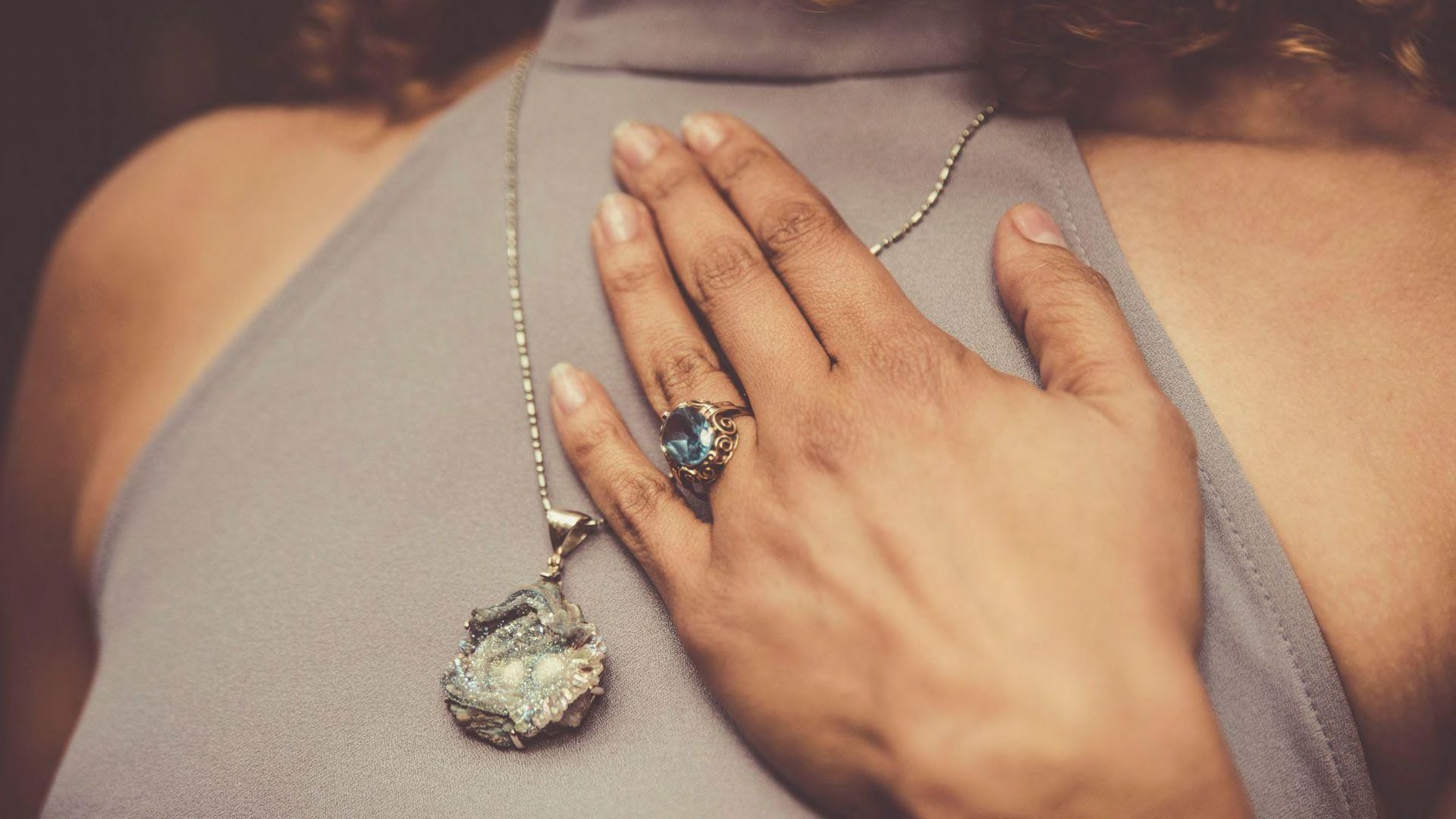 A woman with her hand on her chest wearing a large ring and necklace.