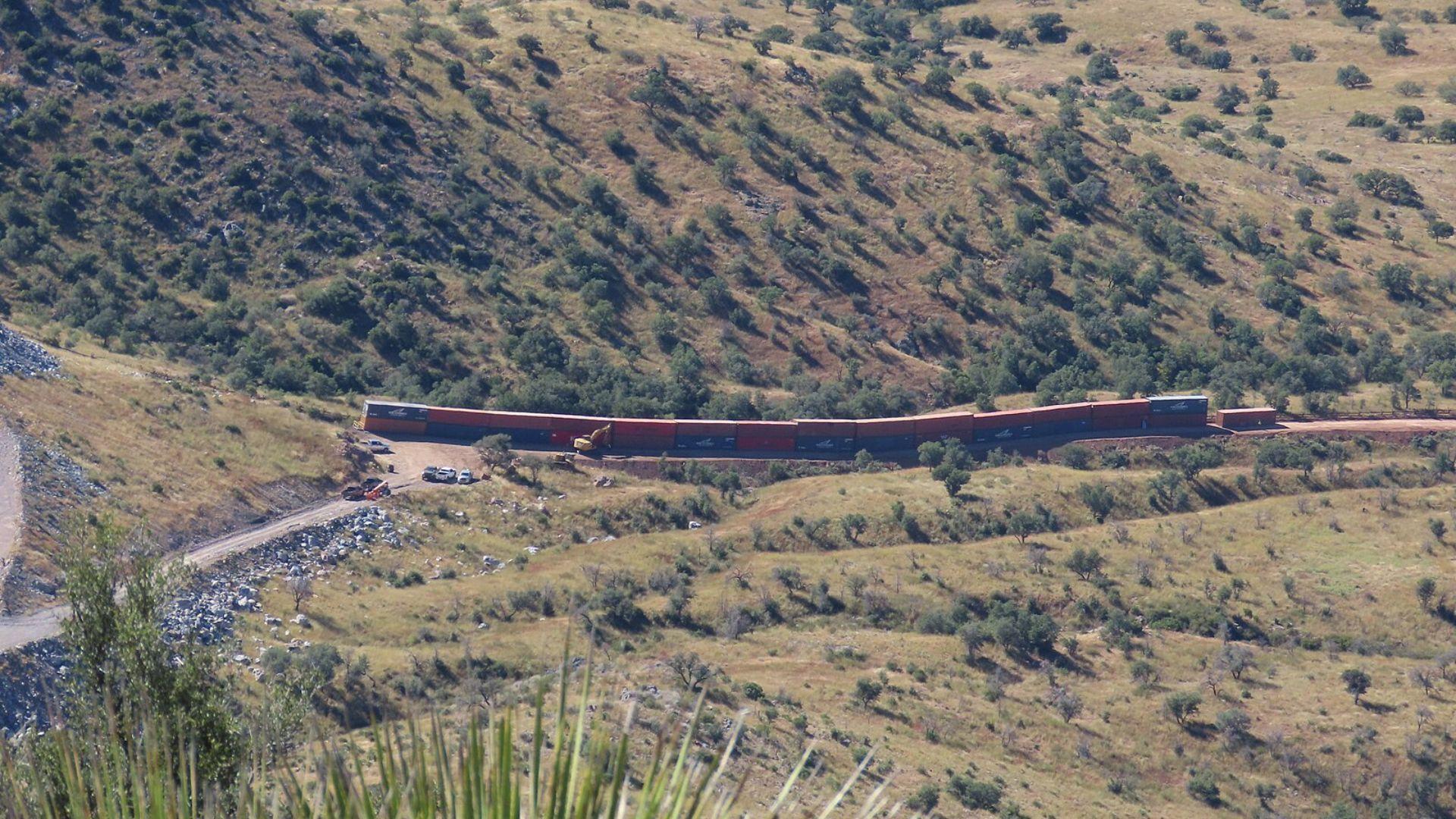 An aerial view of red and blue shipping containers curving around a bend in a mountainous terrain. The containers are surrounded by green scrub-covered hills with scattered trees