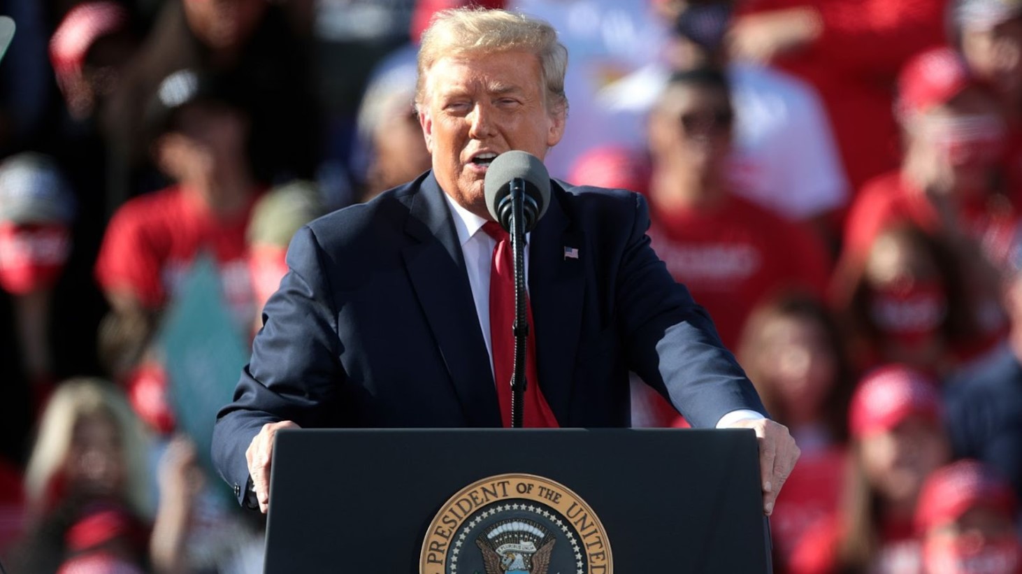 Donald Trump speaking at a podium with the presidential seal, addressing an audience at an outdoor rally, with attendees wearing red caps in the background