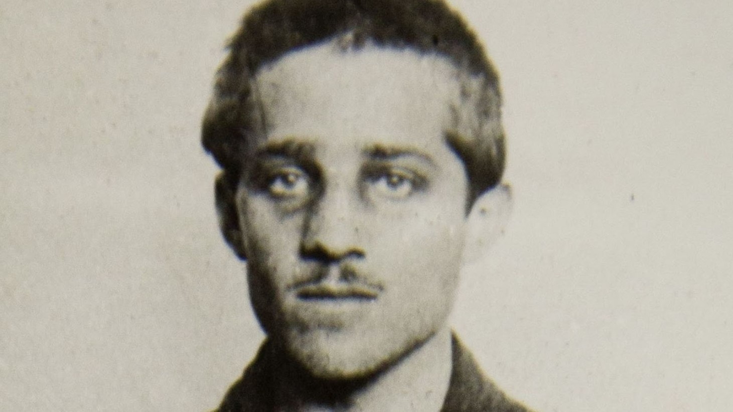 A close-up of an old sepia-toned photograph showing Gavrilo Princip. He is a young man with dark hair and intense eyes, wearing a high-collared shirt