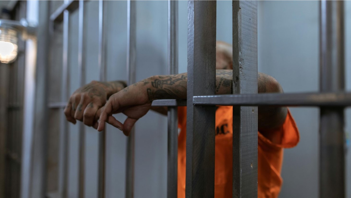 Close-up of tattooed hands gripping the bars of a jail cell, with an orange prison uniform visible in the background