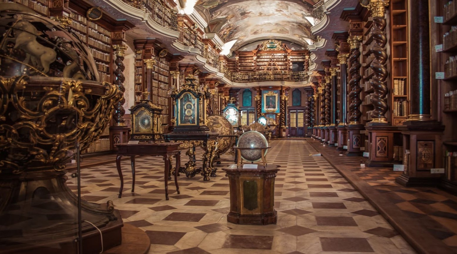An old library. There are bookshelves on either side of the corridor that go up to the ceiling. There are paintings on the ceiling. In the middle of the corridor are two large balls on stands, with some old-fashioned tables.
