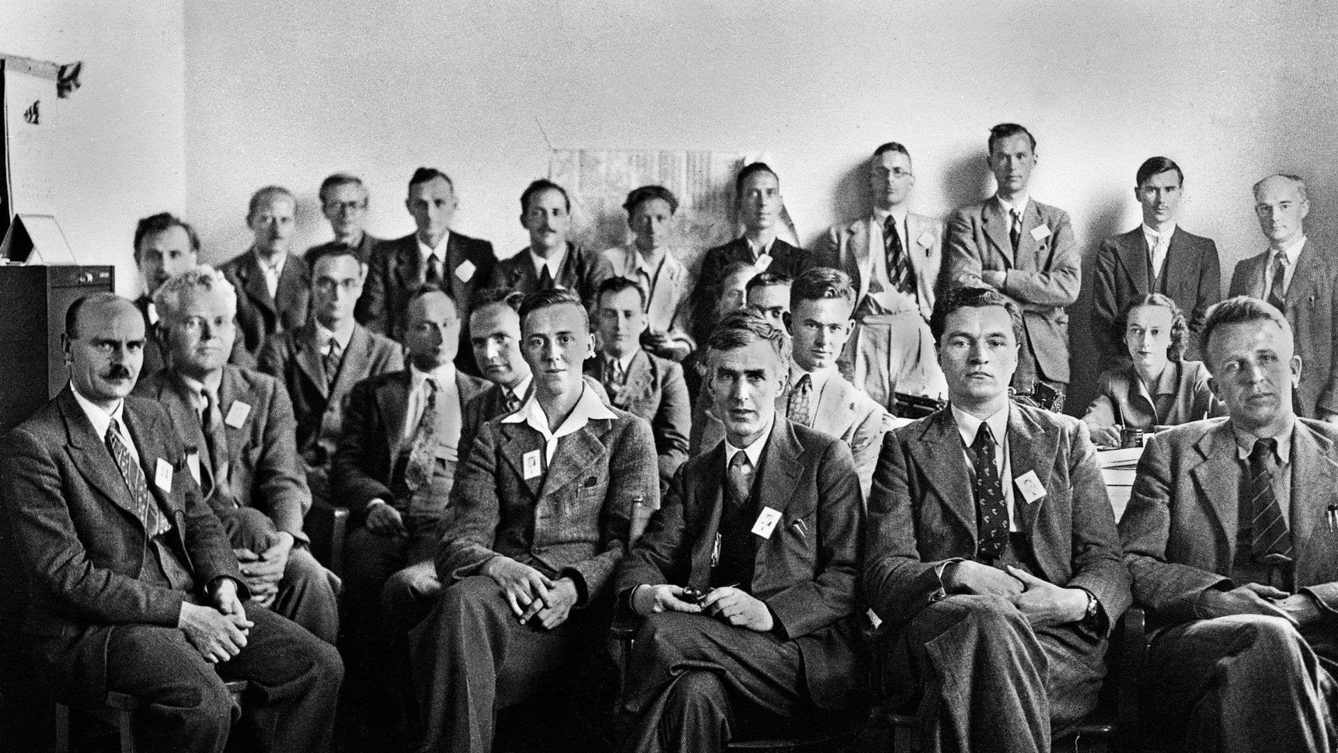 Black-and-white photo of members of the Manhattan Project sitting and standing in a room.