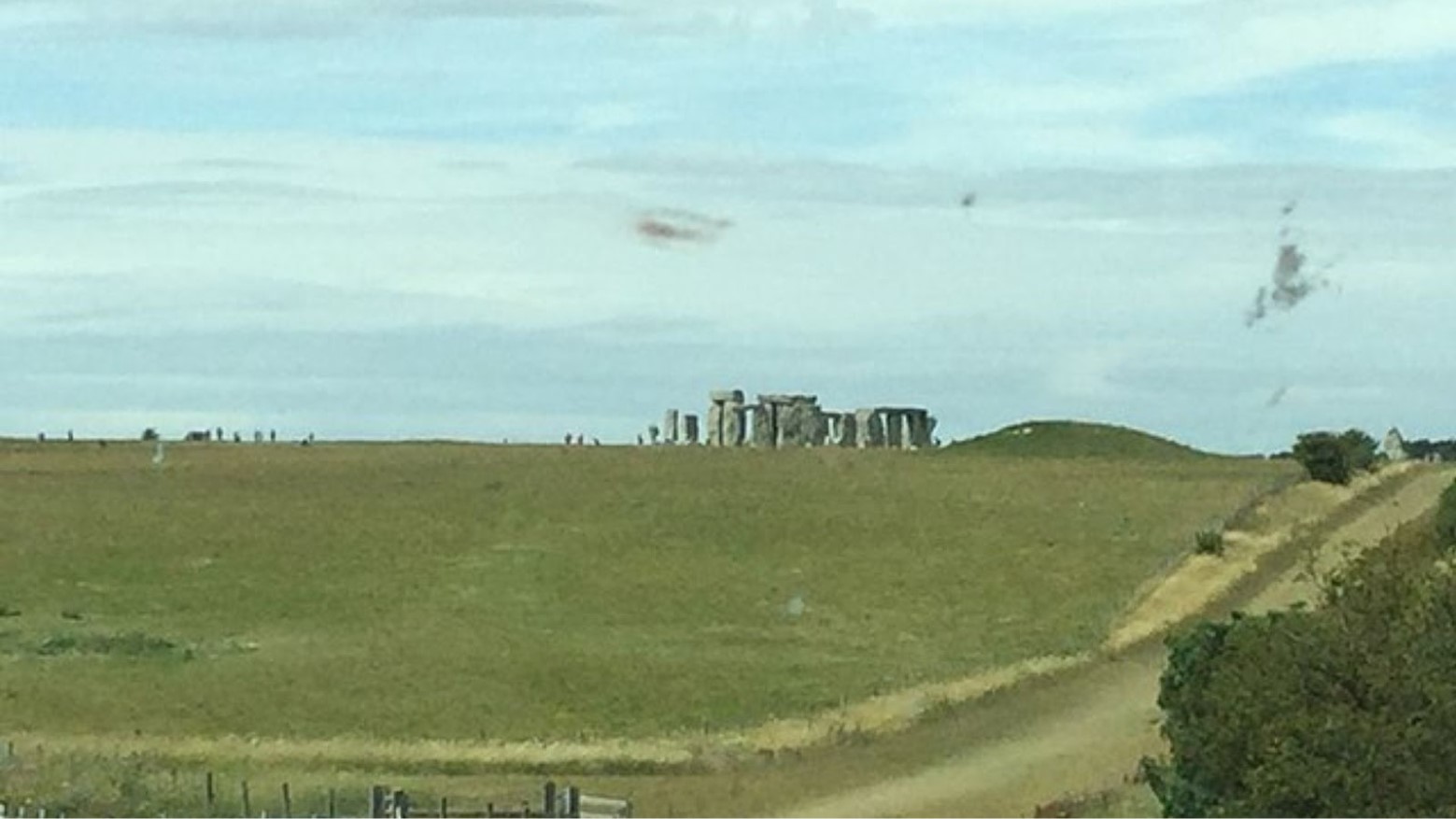 A view of Stonehenge from the road next to it. Stonehenge can be seen in the distance, along with some people going to view it.