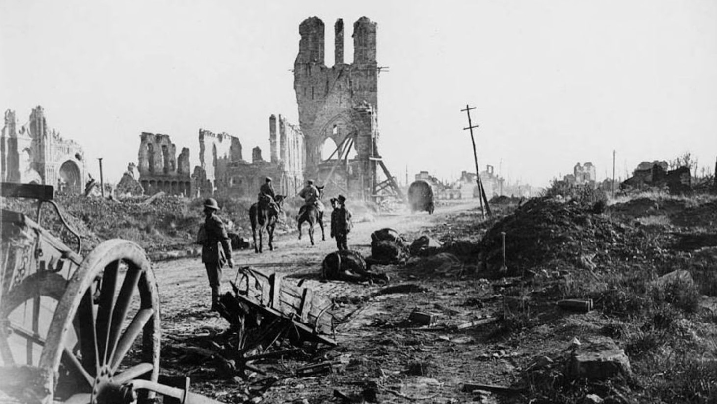 Black and white photograph depicting a devastated World War I battlefield. The foreground shows a broken wagon wheel and debris. Soldiers on horseback and on foot navigate a muddy road amid the ruins