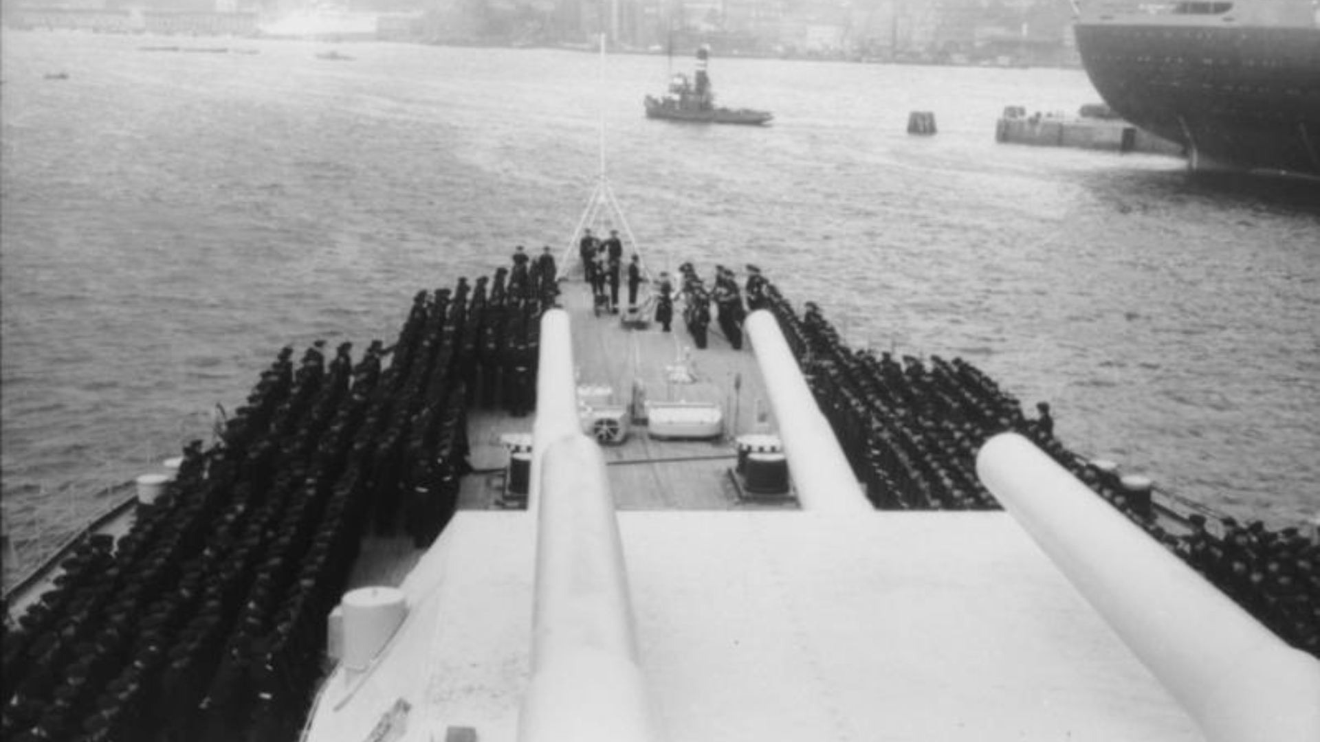Crew members of the Bismarck line up in military formation on the deck. Four of the Bismarck’s gun barrels are in the foreground.