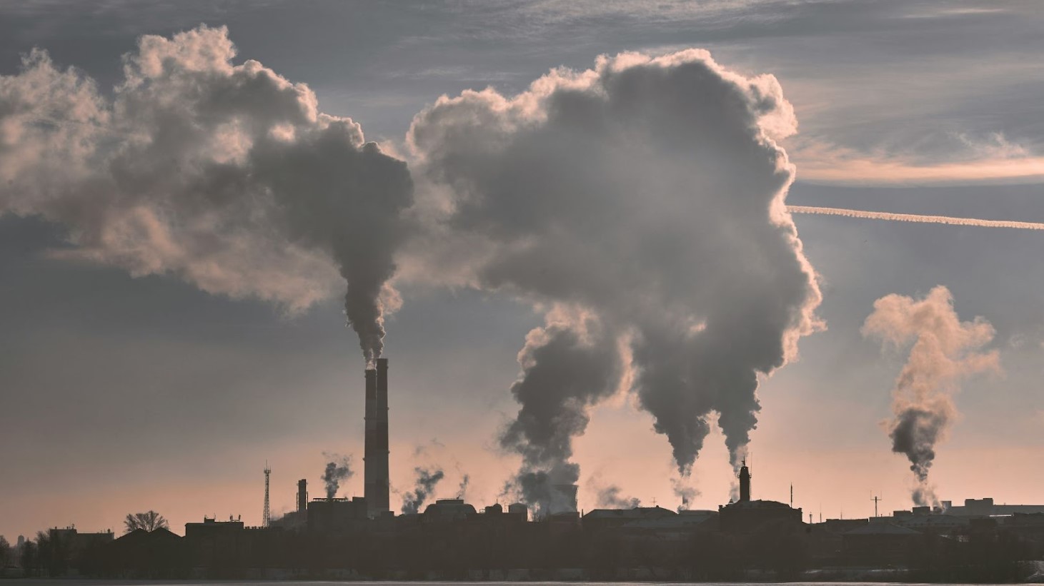 An image of multiple tall industrial chimneys discharging large volumes of dense smoke into the sky at dusk