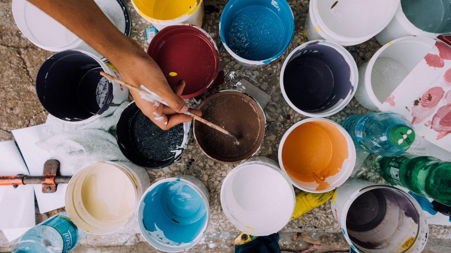A person dipping their paintbrush into a can, with many other colored paints around a table.