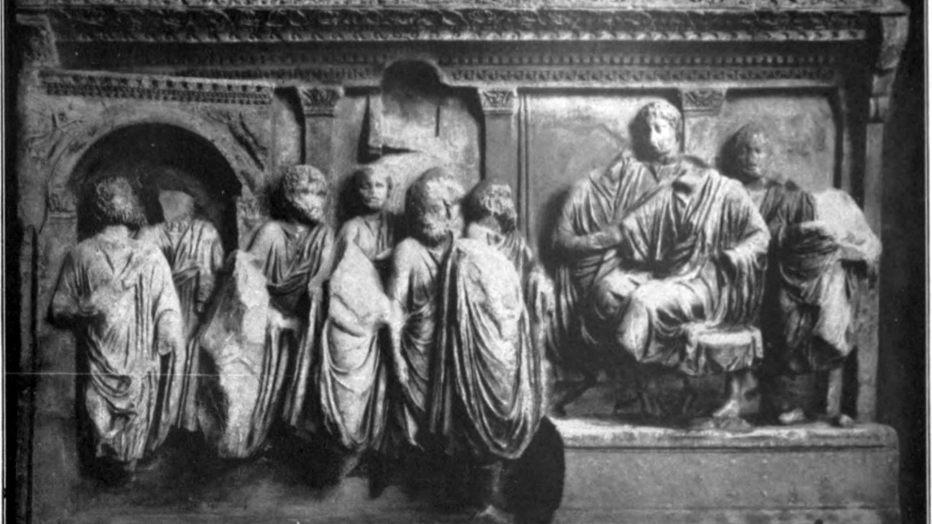Black and white photograph of an ancient Roman bas-relief. The relief depicts a group of figures in Roman attire, some standing and others seated, within an architectural setting that includes columns and an archway