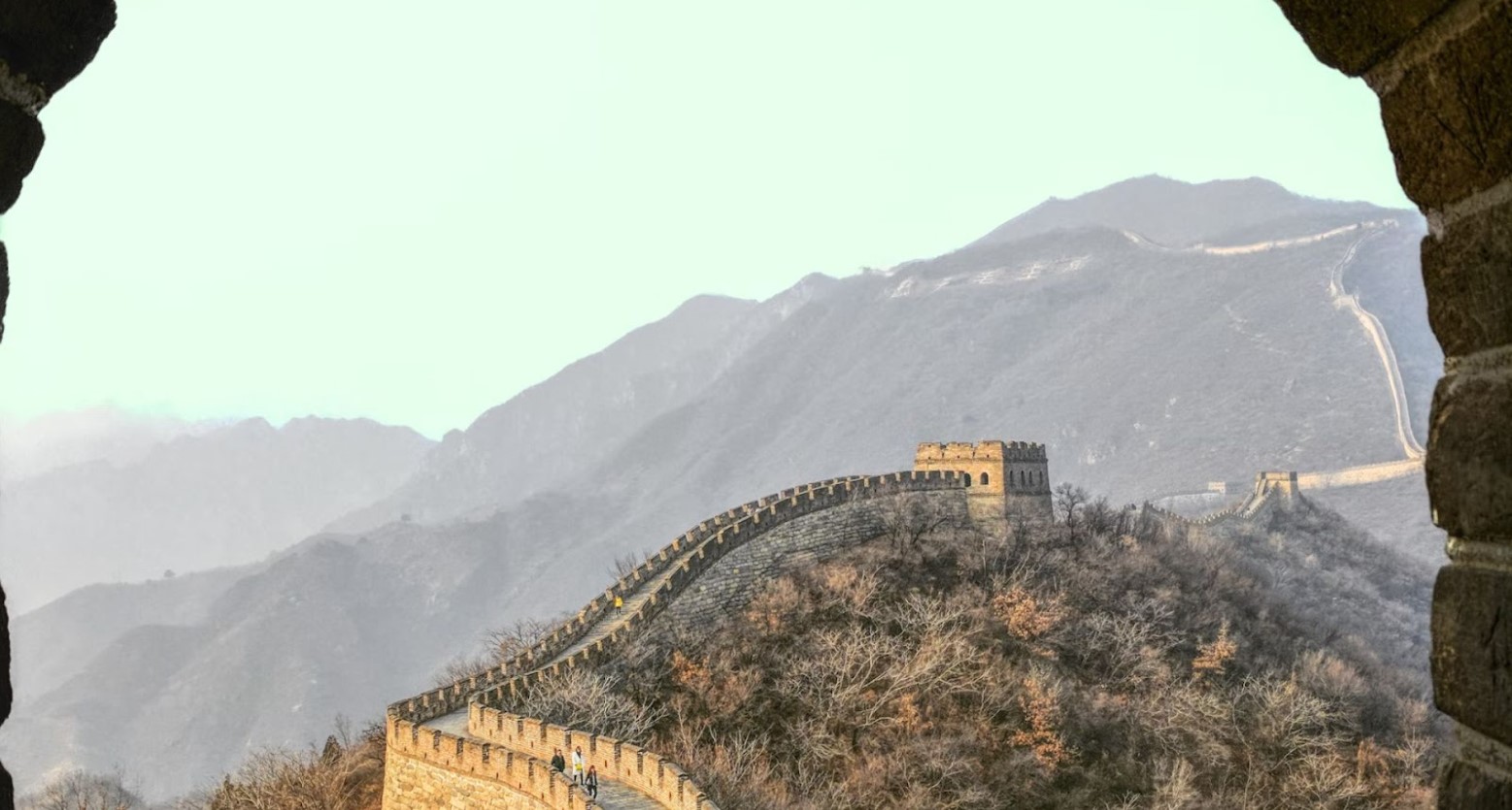 A painting of the Great Wall of China. The painting starts looking through a hole in a brick wall, where you can see the wall go across some hills. Some people are walking along the wall.